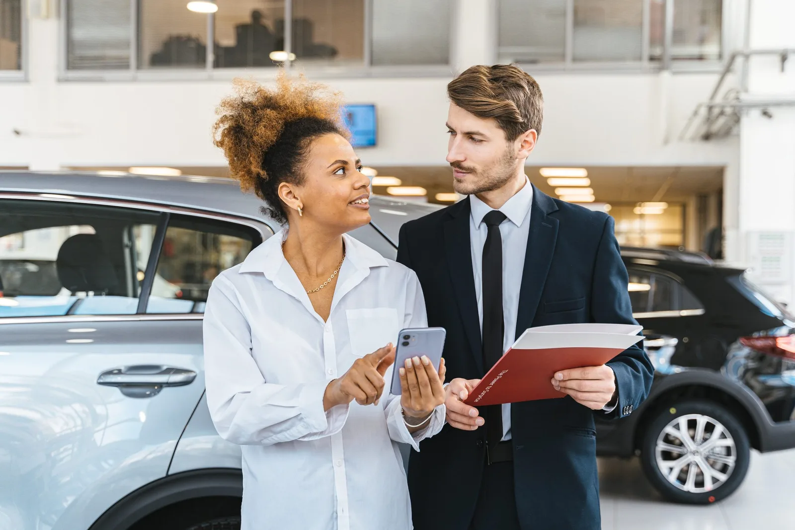 A customer and a salesperson reviewing details on a phone and paperwork inside a car dealership showroom