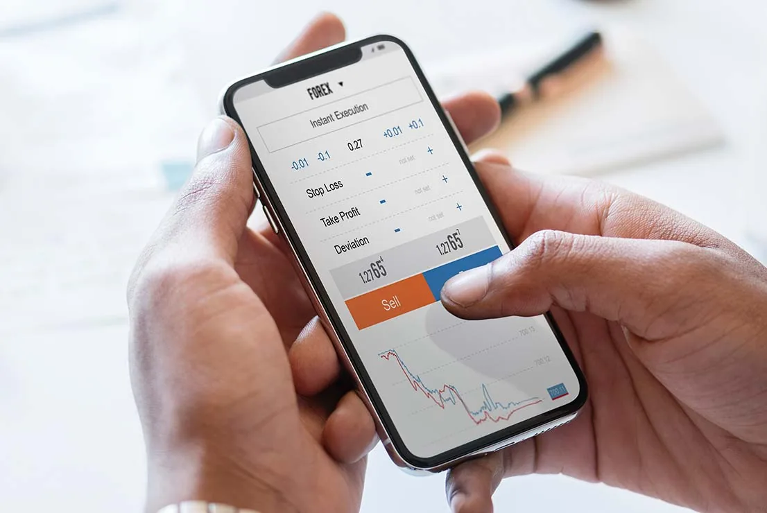 A person using a mobile trading app on a smartphone, held over financial paperwork on a desk