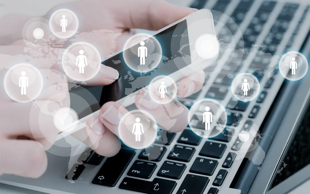 A hand using a smartphone above a laptop keyboard, with glowing bubbles of male and female customer icons floating over a world map